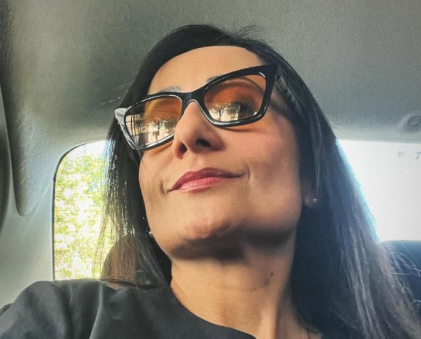 A woman with long hair and thick, black glasses sits in a car.