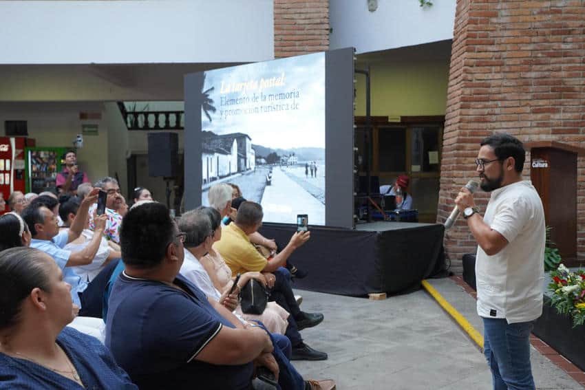 Moisés Hernández López, Puerto Vallarta's official chronicler, gives a lecture on the city's history, with an old photo of the town projected behind him and an audience listening intently.