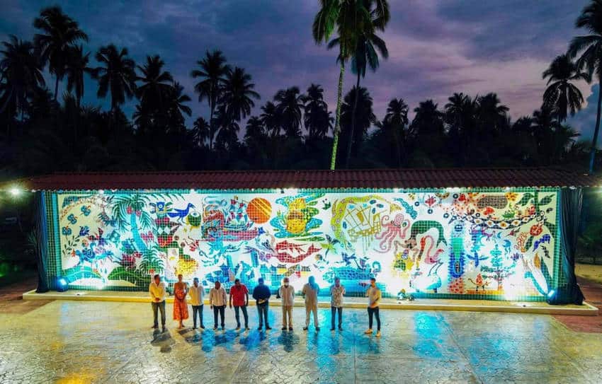 A vibrant, illuminated mosaic mural depicting various colorful and intricate designs, including marine life and abstract patterns, is displayed on a building under a darkening sky. A group of nine people stands in a line facing the mural, admiring it. Palm trees are silhouetted against the twilight sky in the background.