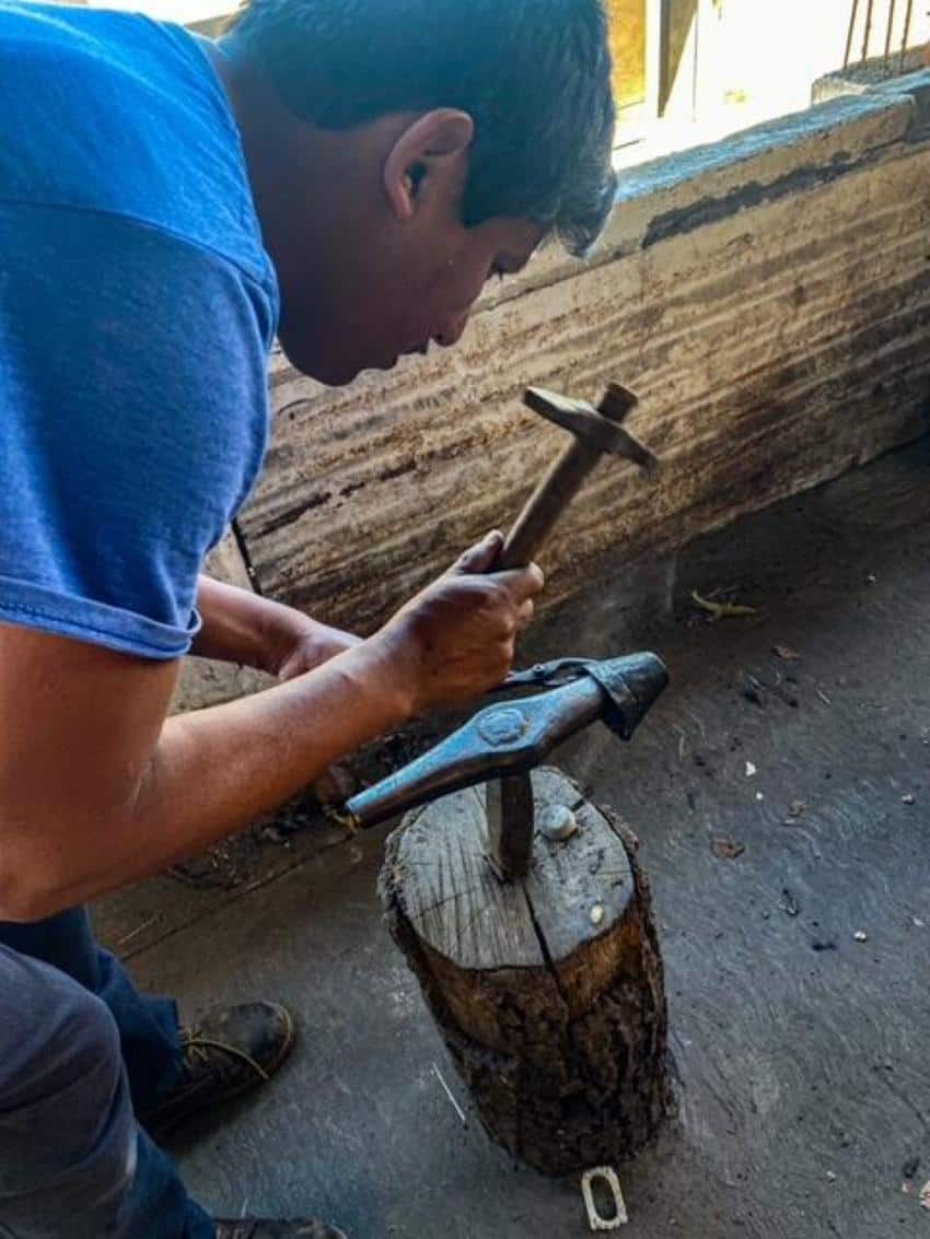 A man leaning down with a small metal hammer to hammer a piece of copper he is holding with a pair of metal tongs