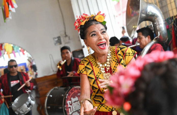 A joyous woman with flowers in her hair, wearing traditional Mexican attire and a necklace with large medallions, sings and dances while a band plays in the background.