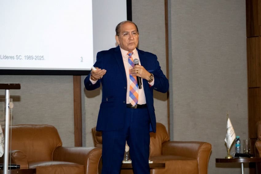 A middle-aged man with short, dark hair and glasses stands in front of a projection screen, actively speaking into a microphone he holds in his left hand. He is wearing a blue suit and a light-colored shirt. His right hand is open in a gesturing motion. Behind him, the screen displays "Líderes SC. 1989-2025." and the number "2". To his right and left, brown leather armchairs are visible, suggesting a conference or presentation setting.