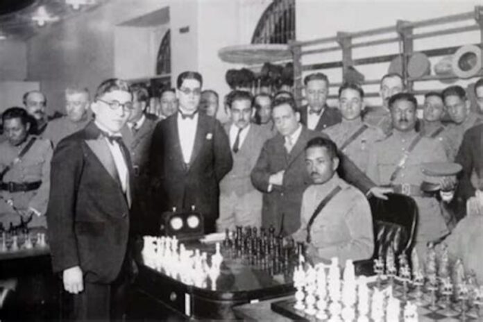 Carlos Torres standing to the left of two men in front of a chess board.