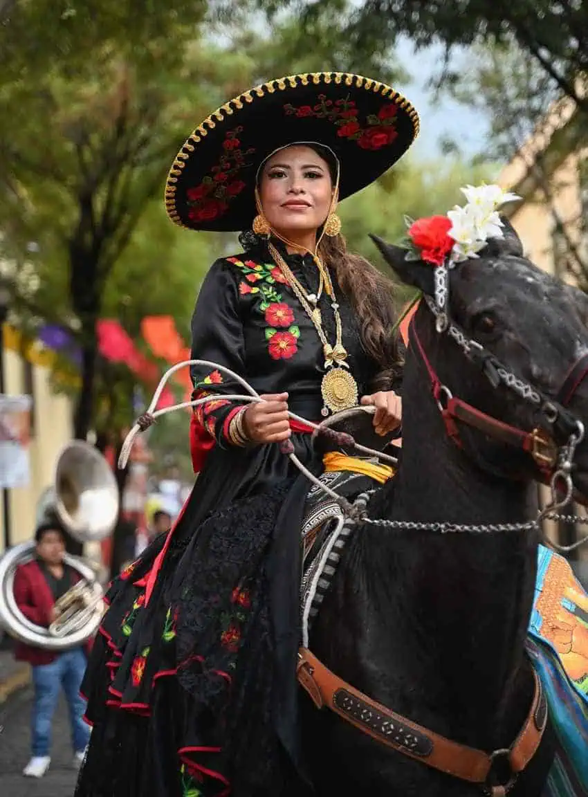 A woman in an elaborately embroidered black charro dress and a wide-brimmed sombrero adorned with flowers, wearing large golden jewelry, sits atop a dark horse and holds reins, looking towards the viewer. Colorful flags are strung across the street in the blurry background.