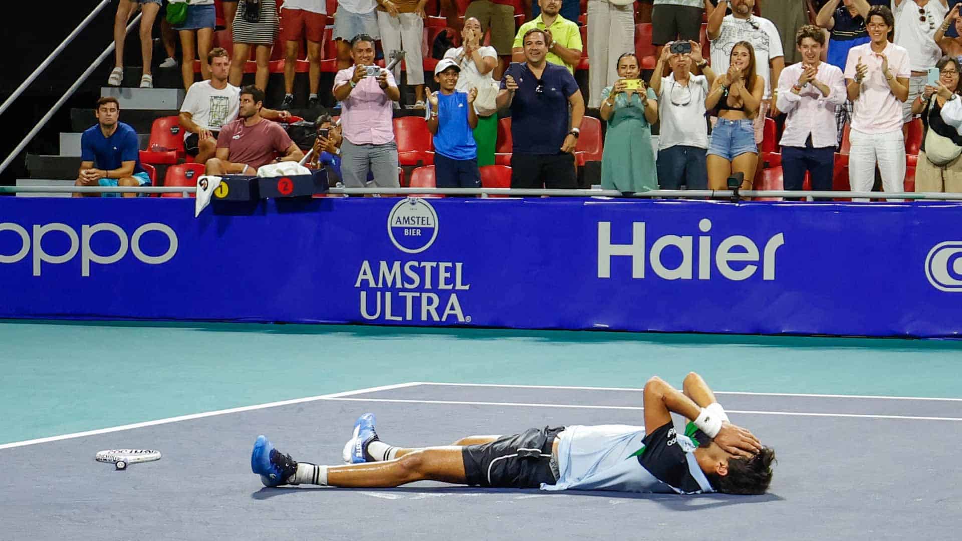 A male tennis player lies on his back on the court, covering his face with his hands in apparent emotion, after a match. Spectators are visible in the stands behind him standing up to applaud or take photos.