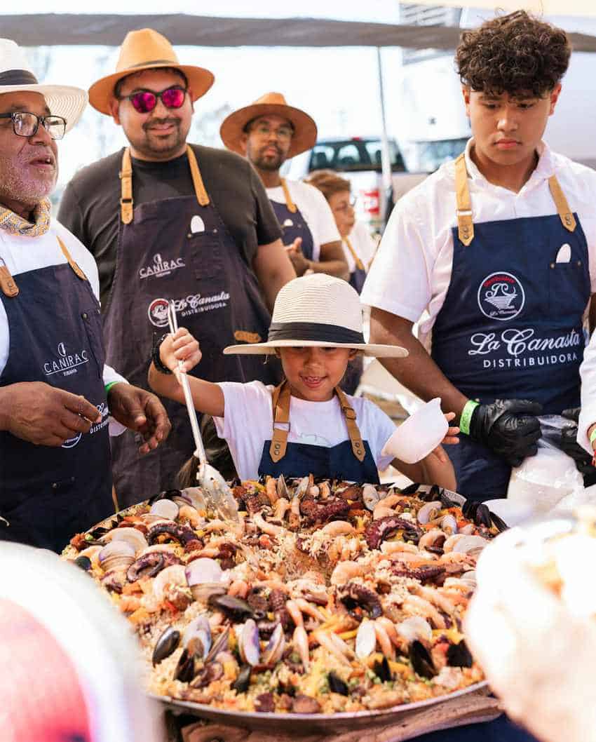 A large group of chefs and cooks, some wearing hats and aprons, are gathered outside in a makeshift kitchen, standing around an enormous paella pan filled with seafood paella. A young boy in a straw hat and apron is enthusiastically stirring the paella, while adults observe and assist at a paella making contest in Baja California, Mexico.
