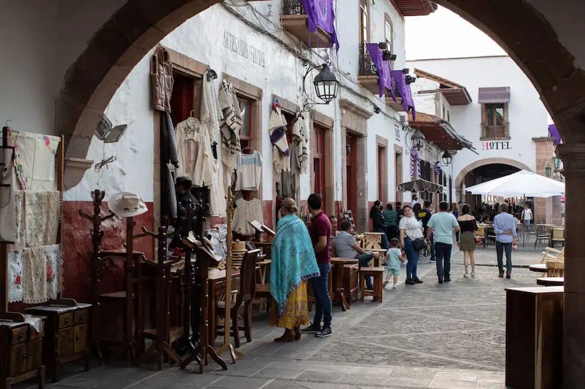 A street scene in Patzcuaro, Mexico, viewed through a stone archway, showing people strolling past shops displaying traditional clothing and crafts. In the background, white buildings with brown trim line the cobblestone street.