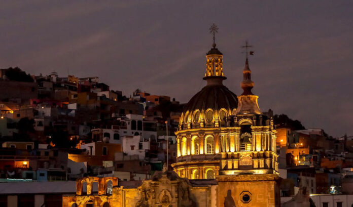 A vibrant night view of Guanajuato, Mexico, featuring a grand, lit-up colonial church with a prominent dome and two spires in the foreground, set against a backdrop of tightly packed, multi-colored houses climbing the hills under a twilight sky.