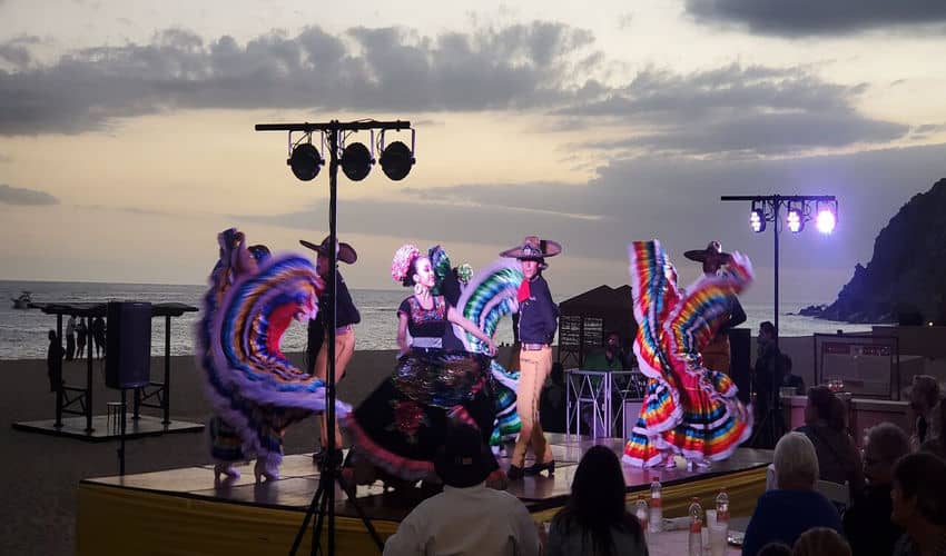 A group of dancers in colorful traditional Mexican dresses and sombreros perform on an outdoor stage on a beach at dusk.
