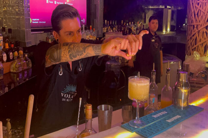 A bartender in a black bar apron focuses on squeezing a lime wedge over a yellow cocktail in a glass on the bar while another bartender looks on in the background.