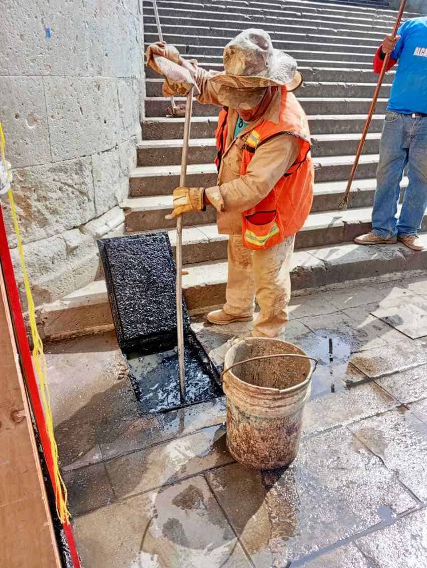 Worker in Mexico in orange construction vest and a camoflauge sunhat epairing urban drainage infrastructure. A man in a tee shirt and jeans holding some kind of pole stands nearby behind him