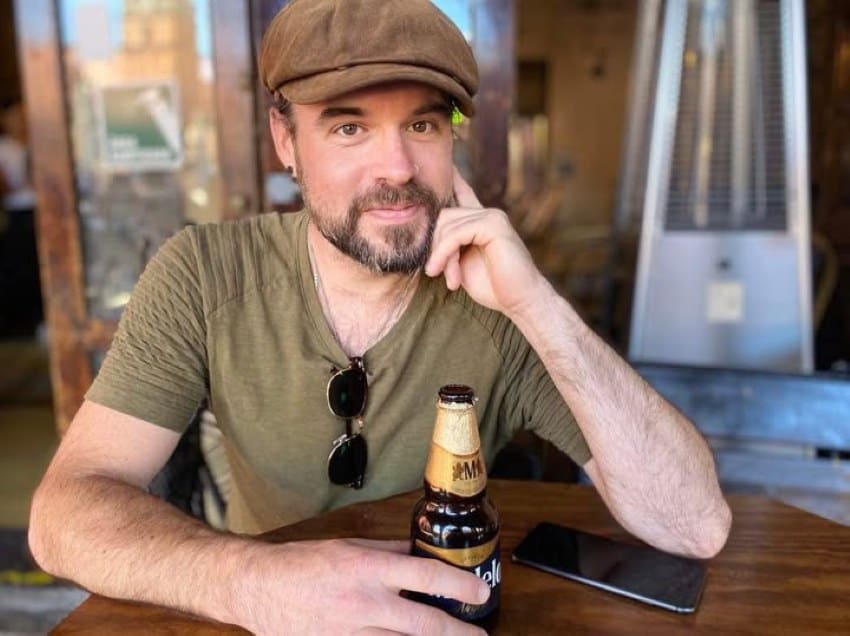 A man in a tee shirt and a newsboy cap poses for a photo while sitting at a round wooden table against a bokeh background.
