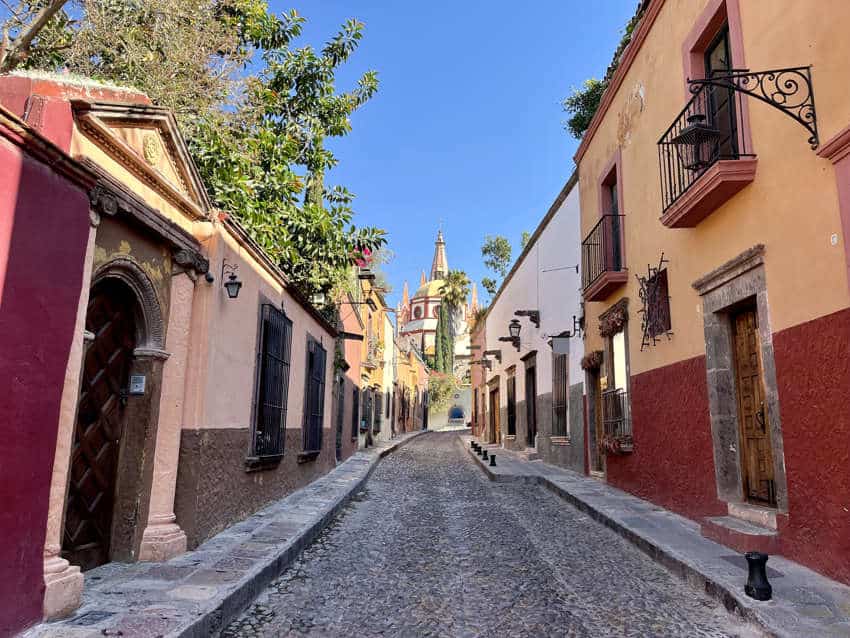 A narrow, uphill cobblestone street in San Miguel de Allende, Mexico, lined with colorful colonial buildings in shades of orange, pink, and white, leading towards a distant church spire under a clear blue sky.