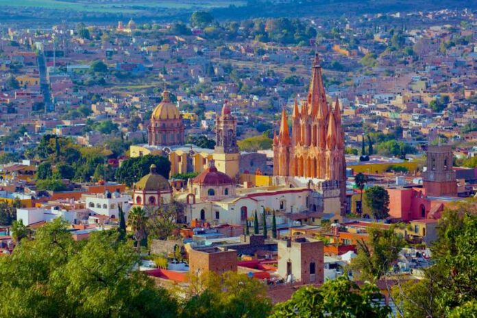 An aerial view of San Miguel de Allende, Mexico, showcasing its dense urban landscape and iconic architectural landmarks under a bright, clear sky. In the foreground, vibrant green trees partially obscure the view. The colonial-era city is characterized by warm, earthy tones like ochre, terracotta, and cream. Prominently featured in the midground is the Parroquia de San Miguel Arcángel, a striking neo-Gothic church with a pinkish-orange facade and towering spires.