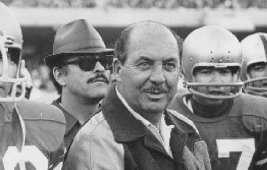 A black and white period photo from the 1940s or 1950s, showing members of a male football team in full uniform and protective helmets stand next to a man in a collared shirt, sunglasses and a pork pie hat and a balding man with a mustache who is in the foreground and the focus of the photo. They are in a football stadium with a crowd in attendance.