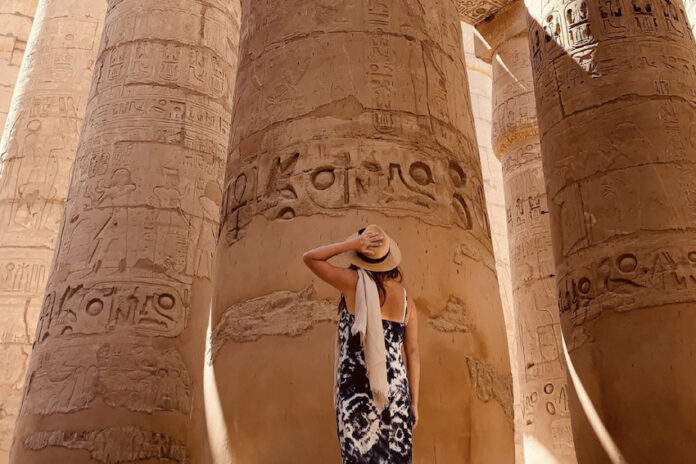 A tanned woman in a sundress and straw hat seen from the back as she looks up at massive pillars of an pre-Hispanic Indigenous structure in Mexico