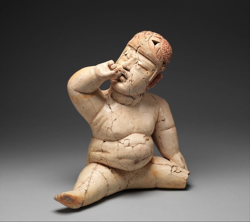 A sculpted Olmec artifact of a fat seated human figure wearing a helmet. Its index finger is up to its mouth as if a baby sucking on its hand.