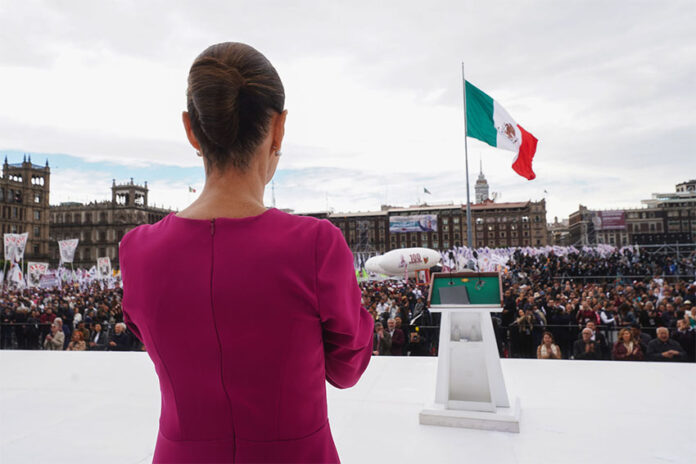 President Claudia Sheinbaum in a pink dress looks out over a crowd in Mexico City Zócalo with the National Palace and a Mexican flag in the background