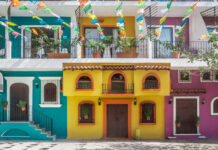What’s on in Puerto Vallarta in August? A street scene in one of Puerto Vallarta's old neighborhoods showcasing the colorful facades of multistory buildings in yellow, purple and bright teal with decorative ironwork on balconies. Overhead, strings of colorful papel picado banners are strung across the street. Potted plants adorn some windows and balconies.