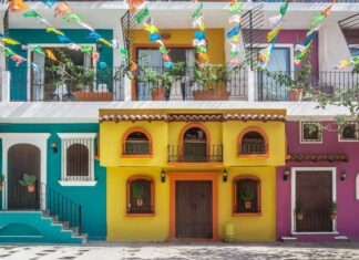 What’s on in Puerto Vallarta in August? A street scene in one of Puerto Vallarta's old neighborhoods showcasing the colorful facades of multistory buildings in yellow, purple and bright teal with decorative ironwork on balconies. Overhead, strings of colorful papel picado banners are strung across the street. Potted plants adorn some windows and balconies.