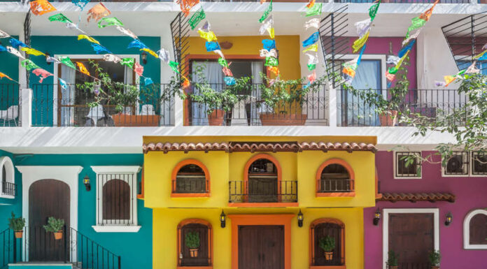 A street scene in one of Puerto Vallarta's old neighborhoods showcasing the colorful facades of multistory buildings in yellow, purple and bright teal with decorative ironwork on balconies. Overhead, strings of colorful papel picado banners are strung across the street. Potted plants adorn some windows and balconies.