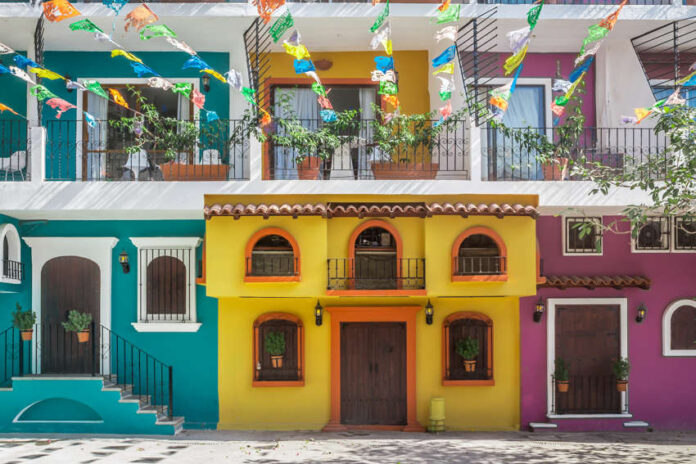 A street scene in one of Puerto Vallarta's old neighborhoods showcasing the colorful facades of multistory buildings in yellow, purple and bright teal with decorative ironwork on balconies. Overhead, strings of colorful papel picado banners are strung across the street. Potted plants adorn some windows and balconies.