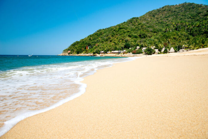 A pristine, empty beach with gentle waves coming in from the Pacific Ocean in Yelapa, Jalisco. In the background is a palm-tree-covered mountain.