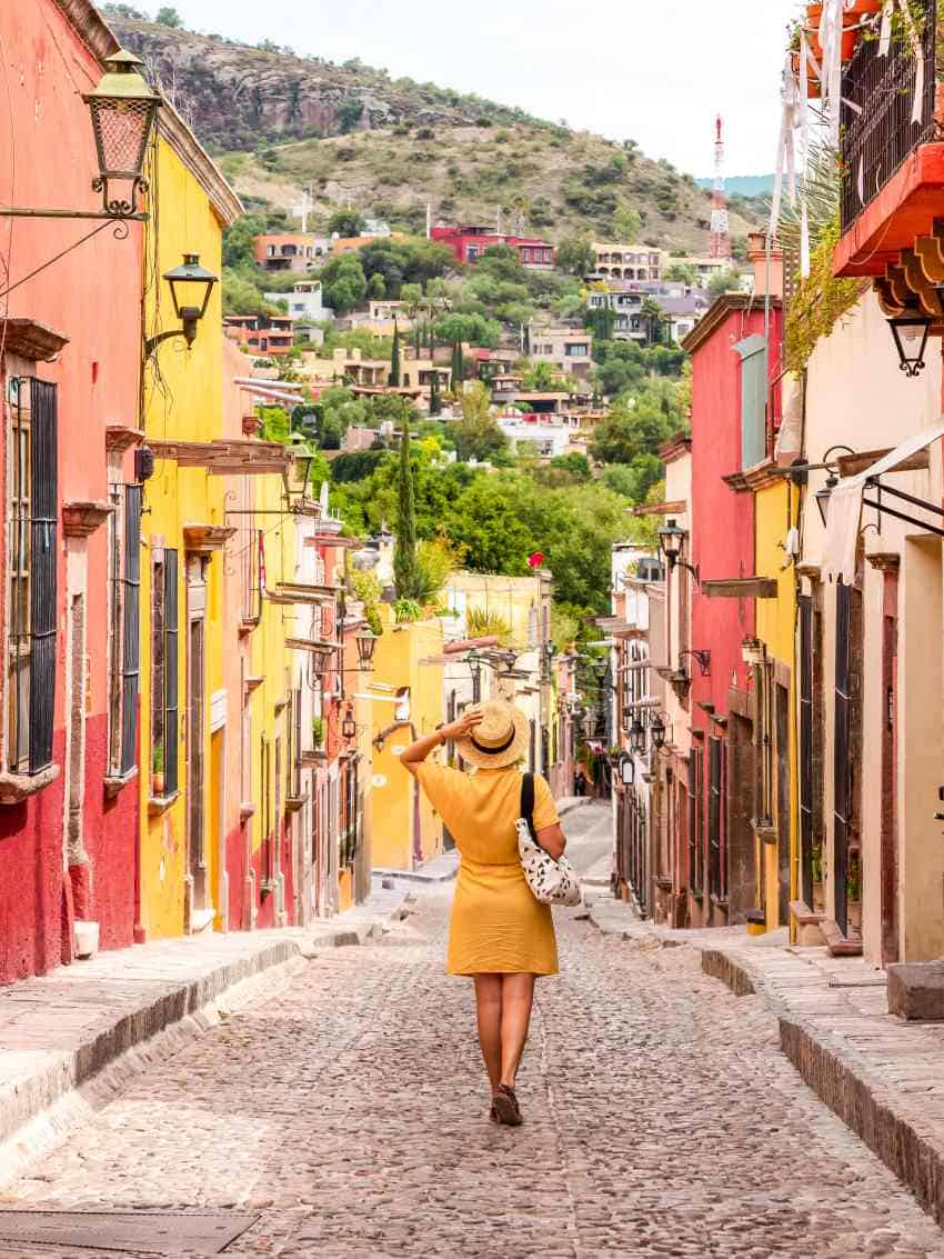 A woman in a yellow dress and a straw hat with a black ribbon band walks down the narrow cobblestoned street with colorful colonial style Mexican buildings on either side of the street in San Miguel de Allende,