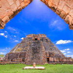 What is Mesoamerica, anyway? A view through a stone archway revealing the ancient, towering Pyramid of the Magician at Uxmal, a Mayan archaeological site, under a blue sky with scattered white clouds. The pyramid is surrounded by green grass and other stone ruins.