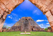 A view through a stone archway revealing the ancient, towering Pyramid of the Magician at Uxmal, a Mayan archaeological site, under a blue sky with scattered white clouds. The pyramid is surrounded by green grass and other stone ruins.
