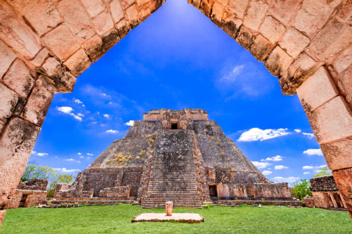 A view through a stone archway revealing the ancient, towering Pyramid of the Magician at Uxmal, a Mayan archaeological site, under a blue sky with scattered white clouds. The pyramid is surrounded by green grass and other stone ruins.