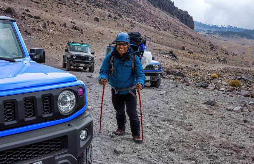 A smiling mountaineer with a backpack and trekking poles stands on a rocky path next to a blue SUV on the lower slopes of Pico de Orizaba, Mexico