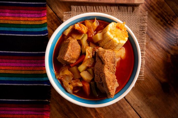 A bowl of mole de olla displayed close up in the center of a table in a food photography style. On the table are also a potholder made of natural fibers and a traditional woven Mexican textile featuring multicolor thin striping.