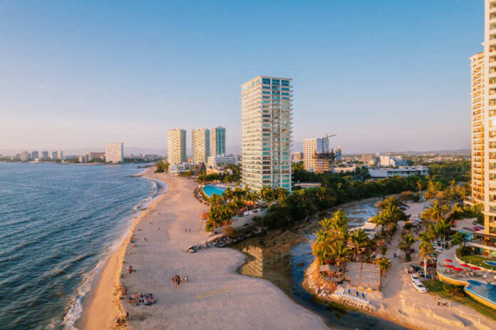 Aerial view of modern high-rise resorts and condominiums lining the sandy beach and coastline of Puerto Vallarta, Mexico, with Banderas Bay stretching into the distance.
