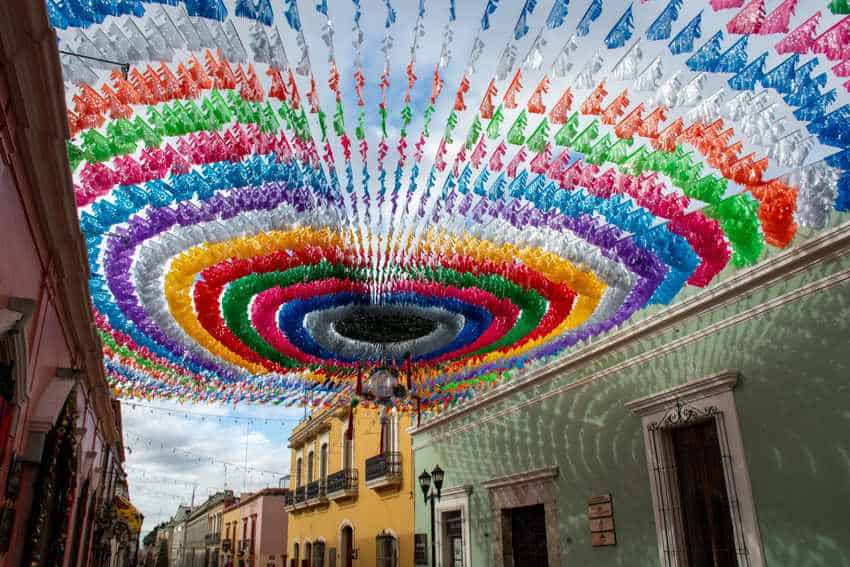 Street in downtown Oaxaca decorated with rows of traditional Mexican colored paper that's tied to buildings on either side of the street, stretching over the street.