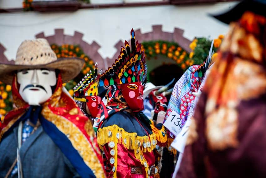 Performers in vibrant traditional costumes and masks, including one with a red face and feathered headdress and another with a straw hat and painted mustache, participating in a cultural celebration or parade in San Luis Potosí, Mexico.