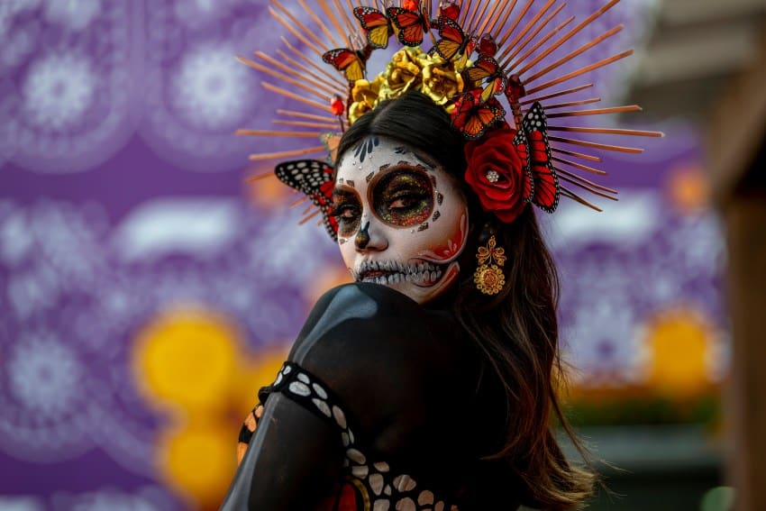 A woman in Day of the Dead Catrina facepaint and a colorful headdress and a dark-green cape, posing sideways and provocatively at the camera.