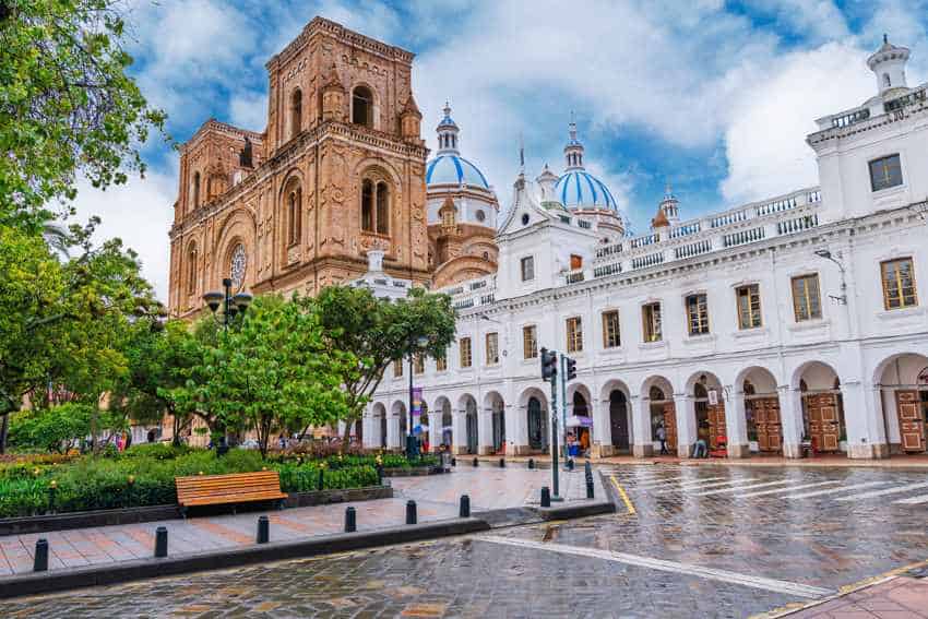 A wide view of the historic city center of the expat retirement haven of Cuenca, Ecuador. Visible is the Nueva Catedral and its iconic blue domes and a grand stone facade, alongside white colonial buildings with arches, a green park with a bench, and a wet cobblestone street after rain, under a partly cloudy sky.