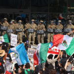 Protesters with Mexican flags and signs stand in front of a wall of California National Guard troops