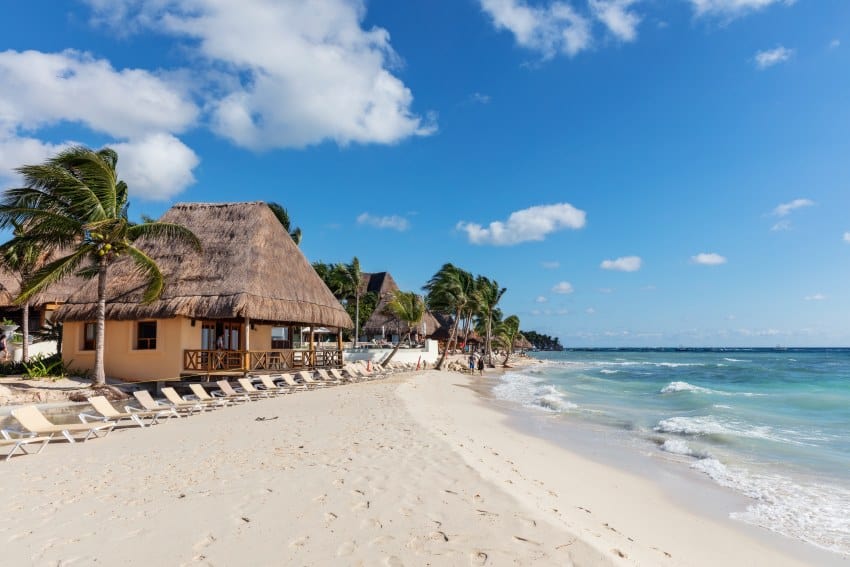 A bungalow near the shore of a beach in Playa del Carmen, Mexico. In front of it are white beach chairs facing the mild waves of the ocean.