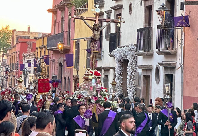 Men in black suits parade down a San Miguel de Allende street holding a figure of Christ on the cross over their heads.