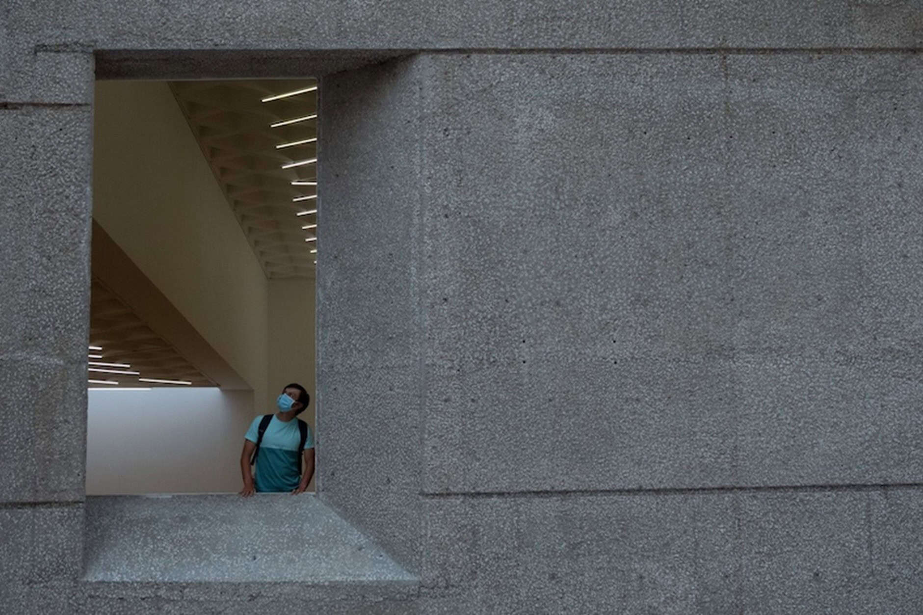 A man wearing a light blue face mask and a backpack looks up and to the right, framed by a concrete window opening. The window is part of a large, textured concrete wall, and inside the building, angled light fixtures are visible on the ceiling.