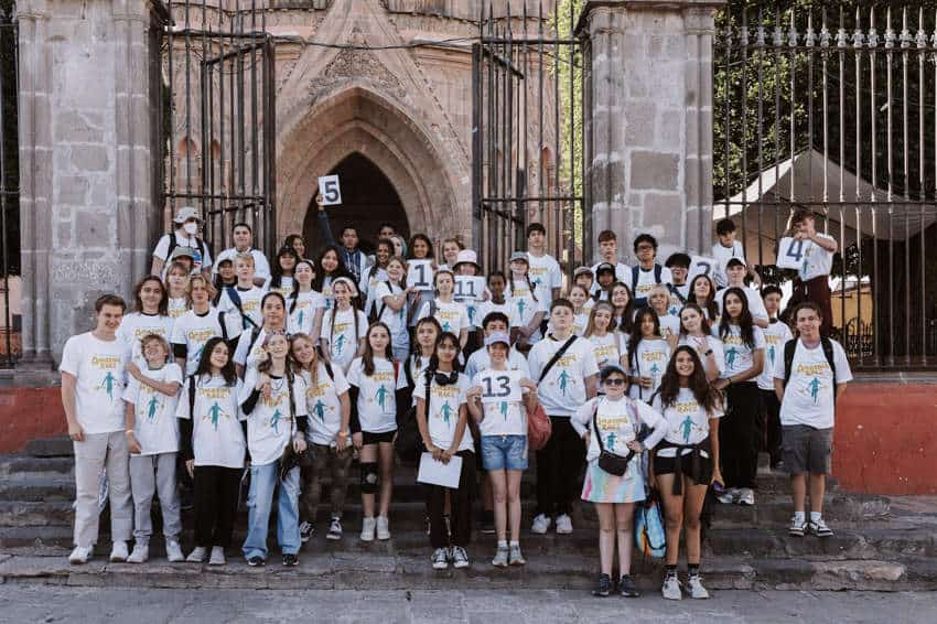 A large group of teenagers and young adults are posing for a group photograph on the stone steps in front of an ornate, historic church in San Miguel de Allende after the city's annual The Amazing Race. Most are wearing matching white t-shirts with a logo, and several are holding up signs with numbers that appear to be their racer placards.