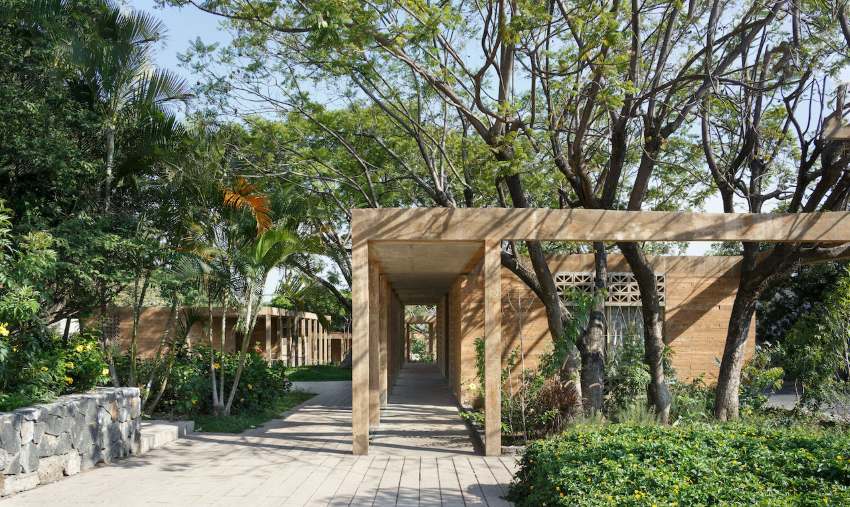 A rectangular stone building with a brick pathway, surrounded by tall trees bordered by a stone wall