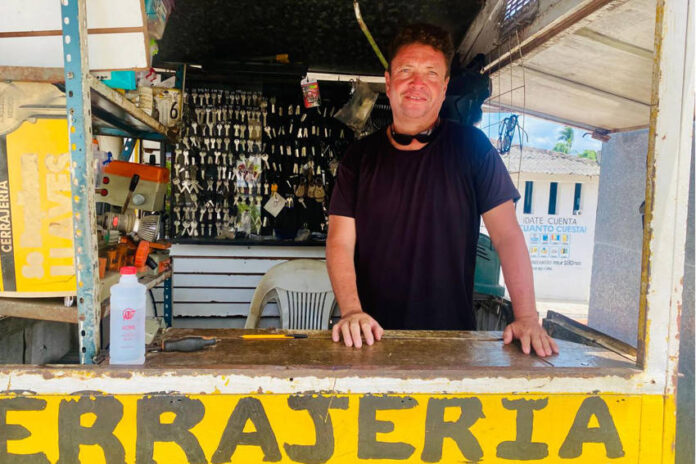 A man with a smiling expression and sunglasses pushed up on his head stands behind a counter at an outdoor key-making stand. Behind him, numerous keys are hanging, and the word 