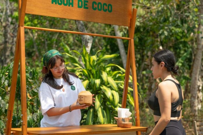 Young woman standing at a wooden restaurant serving booth handing over a coconut shell glass of coconut water to a young female customer on the other side of the booth, which is located outside in a verdant background of trees.