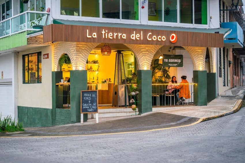 Outer facade of La Tierra del Coco cafe. It has three green arches, is located on a street corner. It has warm yellow lighting inside and you can see a couple of women sitting together inside at a table overlooking the street.