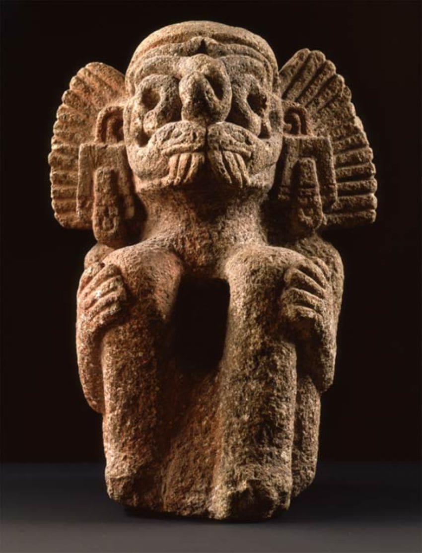 A 40 cm high stone carved scuplture of a humanoid god figure sitting with his knees up and his hands on his knees. He is staring upward. His eyes are made of carved serpents curved in an oval. He wears a carved headdress made to look like it's made of feathers.