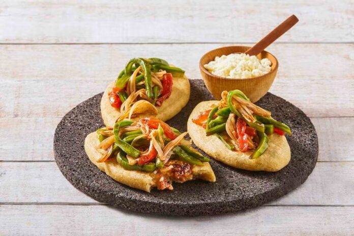 Three tlacoyos topped with peppers and onions, sitting on a serving platter made of volcanic rock. Also on this platter is a small wooden bowl of white rice with a matching wooden spoon for serving.