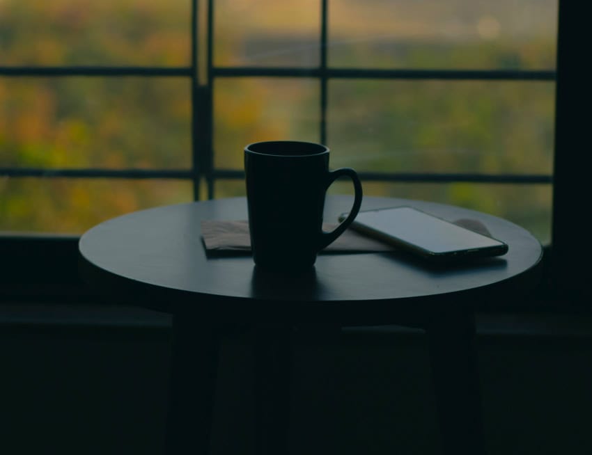 A dark silhouette of a coffee mug and a smartphone on a round table, set against a blurred window showing green foliage and warm, soft light.